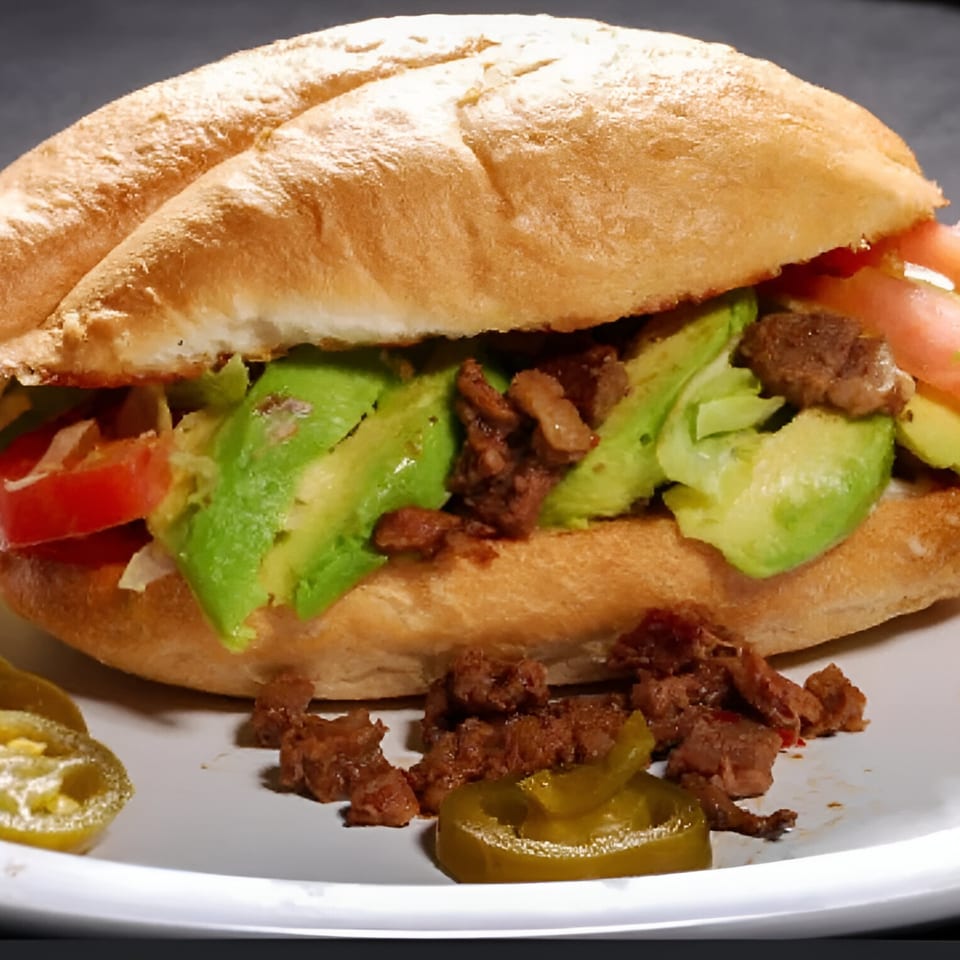 A large torta sandwich filled with meat, avocado, lettuce, and tomato, served with jalapeños.